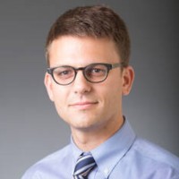 Portrait of a researcher wearing glasses and a blue shirt, likely involved in cancer research.